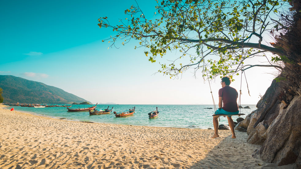 A man sits in a tree swing looking out over boats on a beach in Thailand